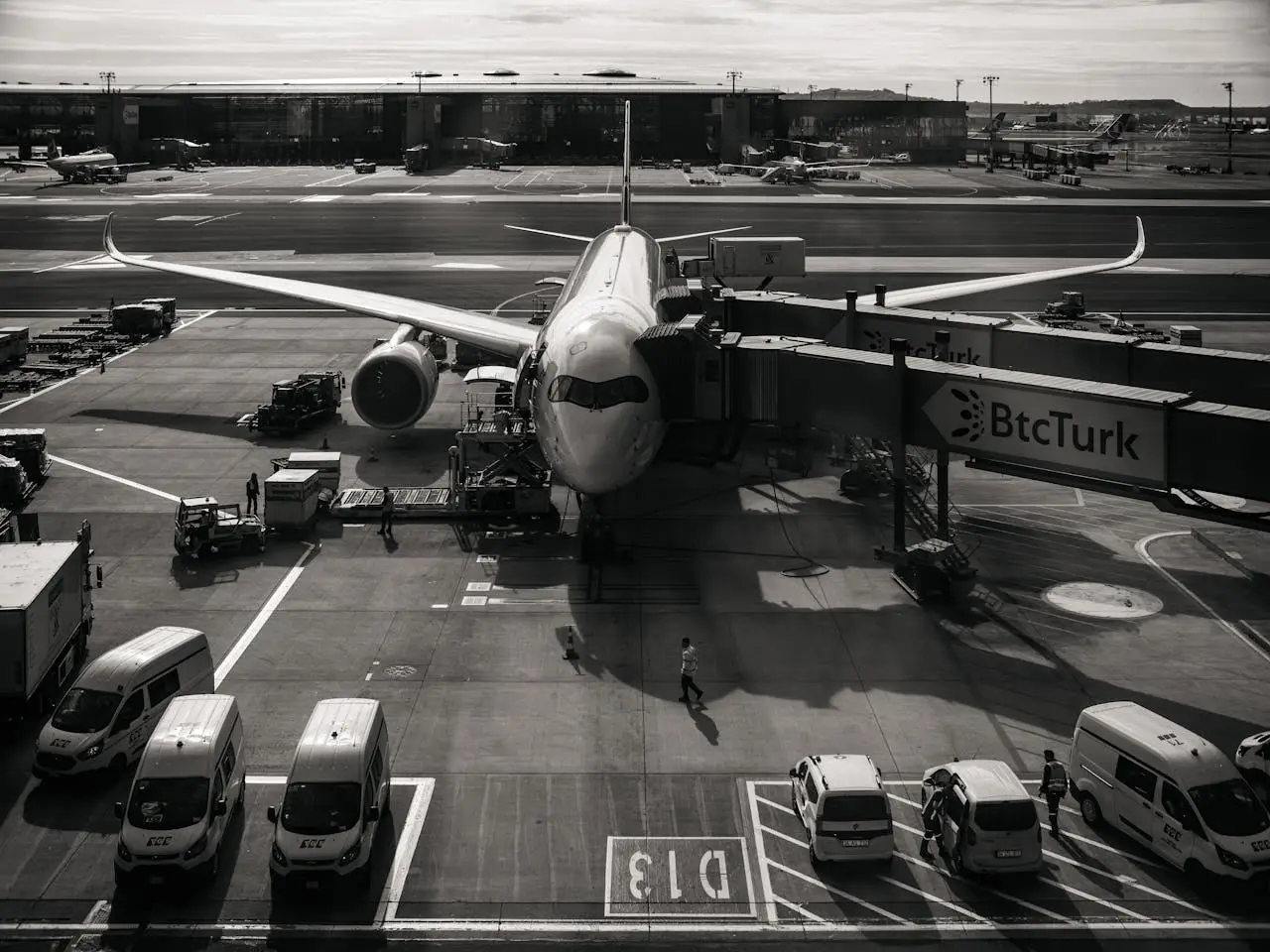 Services Monochrome image of airplane at airport gate with vehicles and jetway during the day.